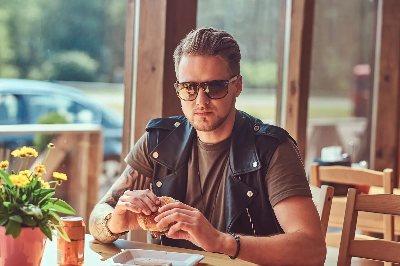 A hipster with a fashionable hairstyle and beard sits at a table, having decided to dine at a roadside cafe, and is eating a hamburger.