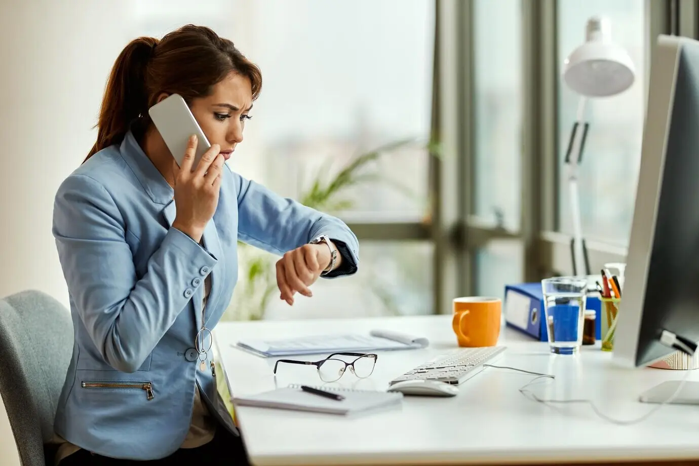 A distraught businesswoman talks on the phone in the office while checking her wristwatch for the time.