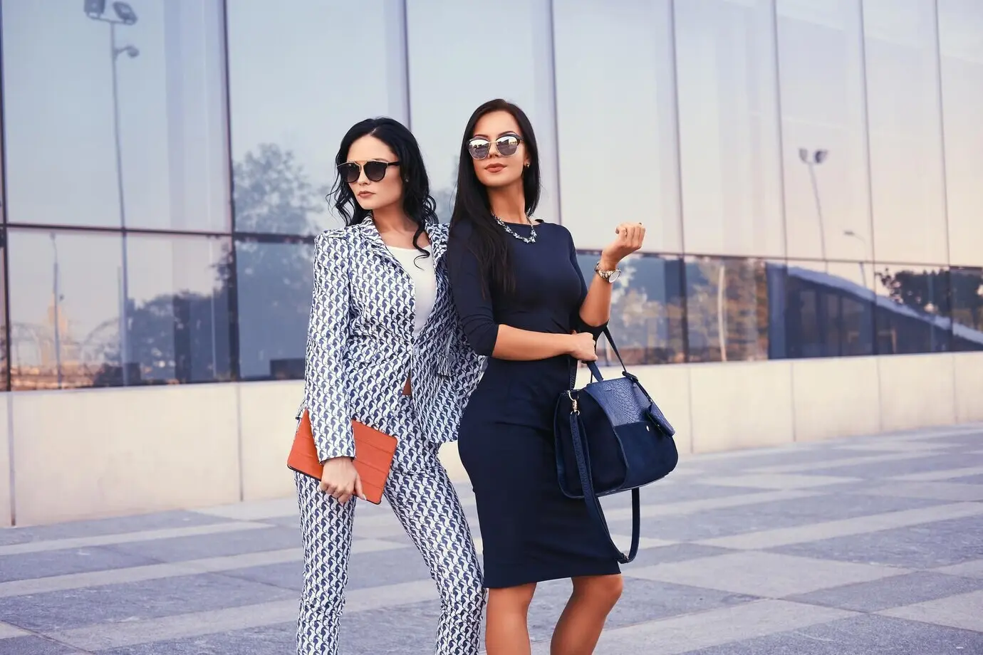 Portrait of two businesswomen in stylish formal attire, standing downtown with a skyscraper in the background.