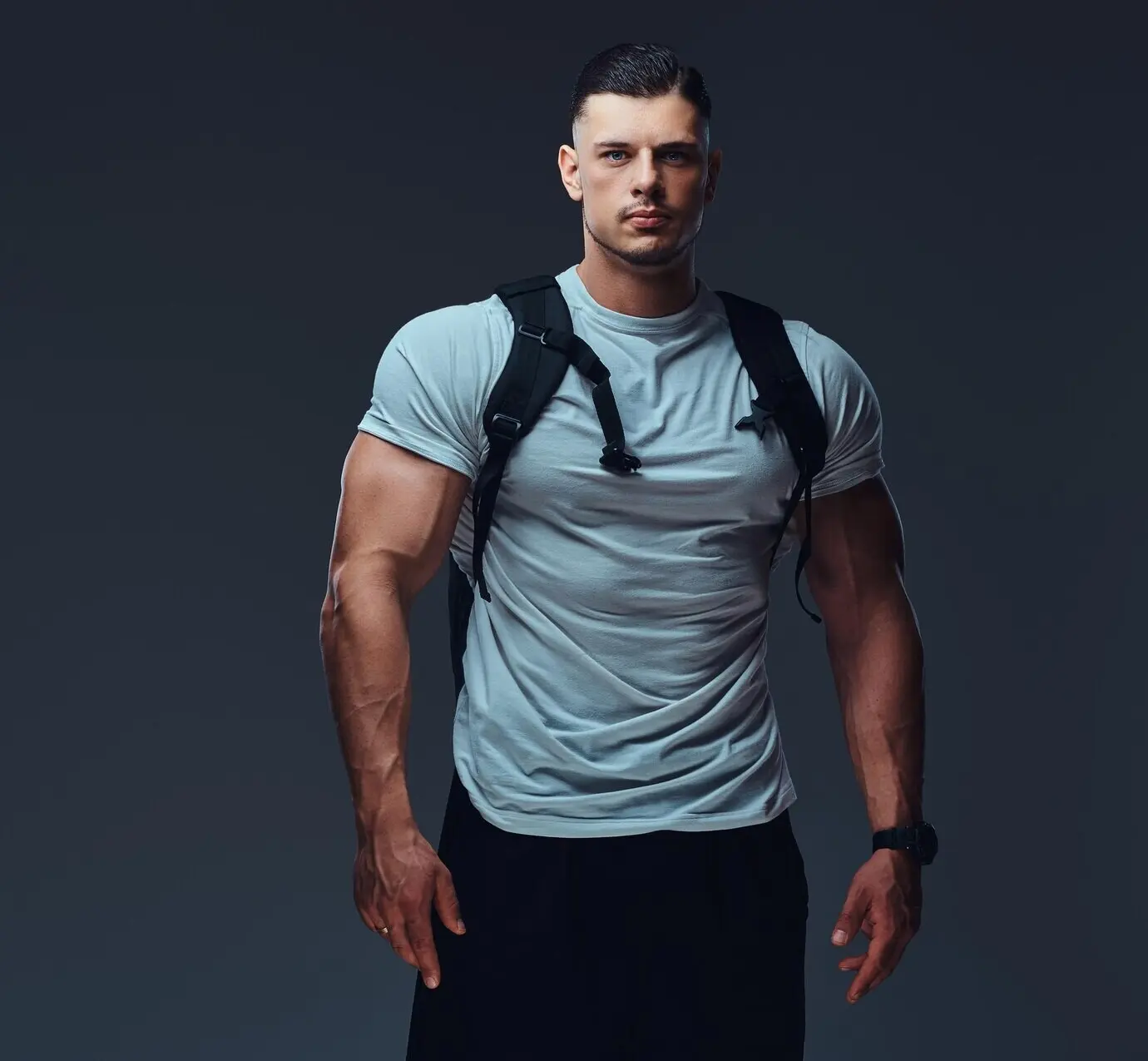 A well-muscled, good-looking bodybuilder in athletic wear, carrying a backpack, strikes a pose before a gray backdrop.