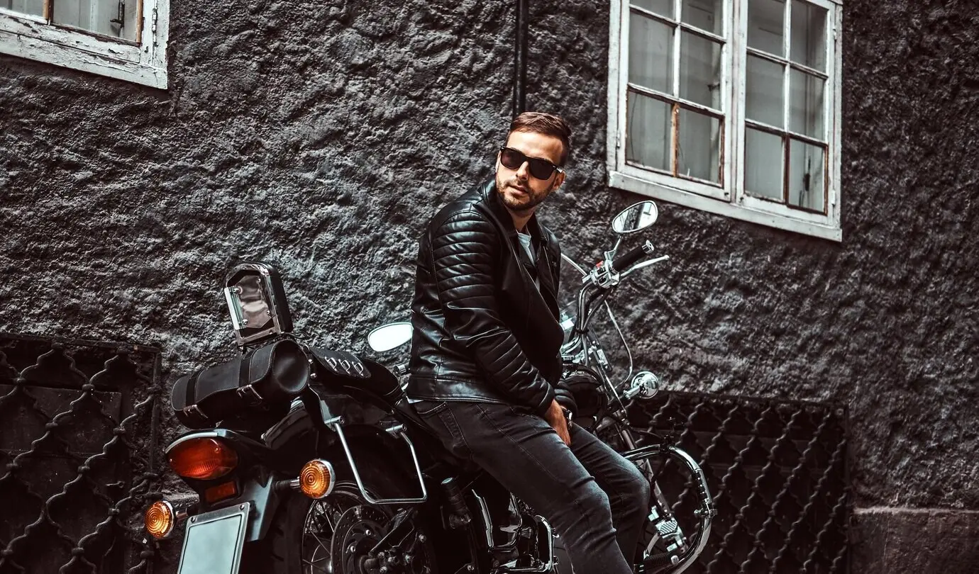 A stylish young biker in sunglasses, wearing a black leather jacket and jeans, seated on his vintage motorcycle on an old European street.