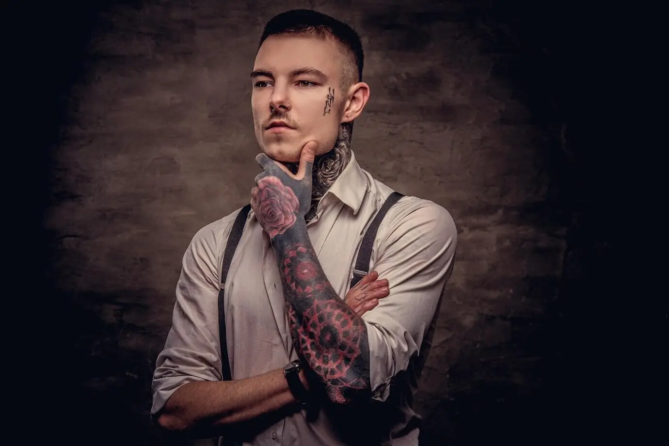 Close-up portrait of a pensive young, old-fashioned tattooed guy in a white shirt and suspenders, with his hand on his chin, isolated against a dark background.
