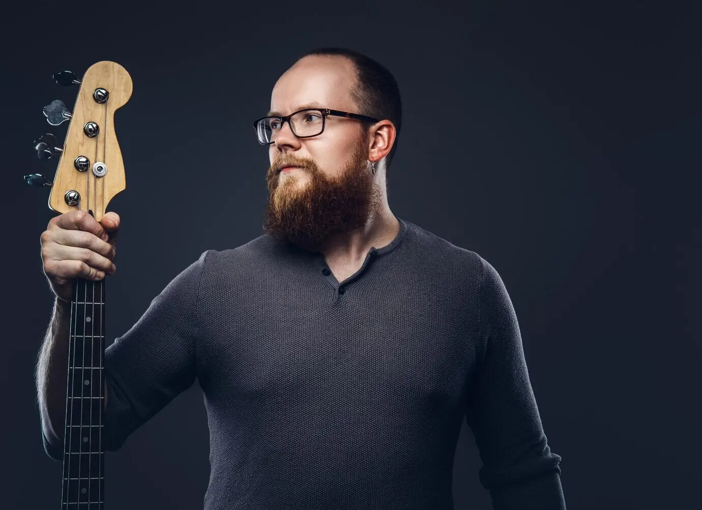 A red-haired, bearded male musician wearing glasses and a gray T-shirt holds an electric guitar, isolated against a dark, textured background.