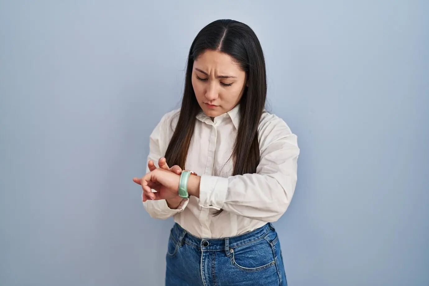 A young Latin woman stands against a blue background, checking the time on her wristwatch, relaxed and confident.